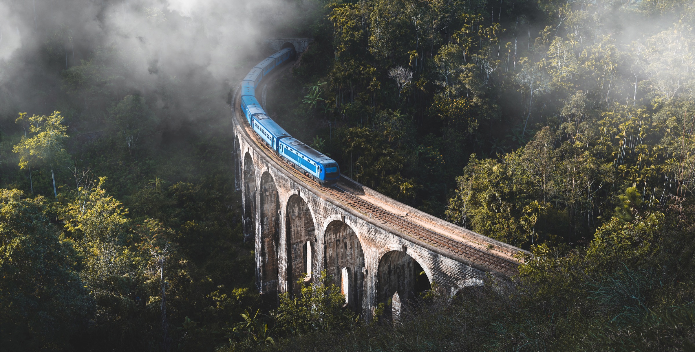 A blue train crossing Nine Arch Bridge through misty hills