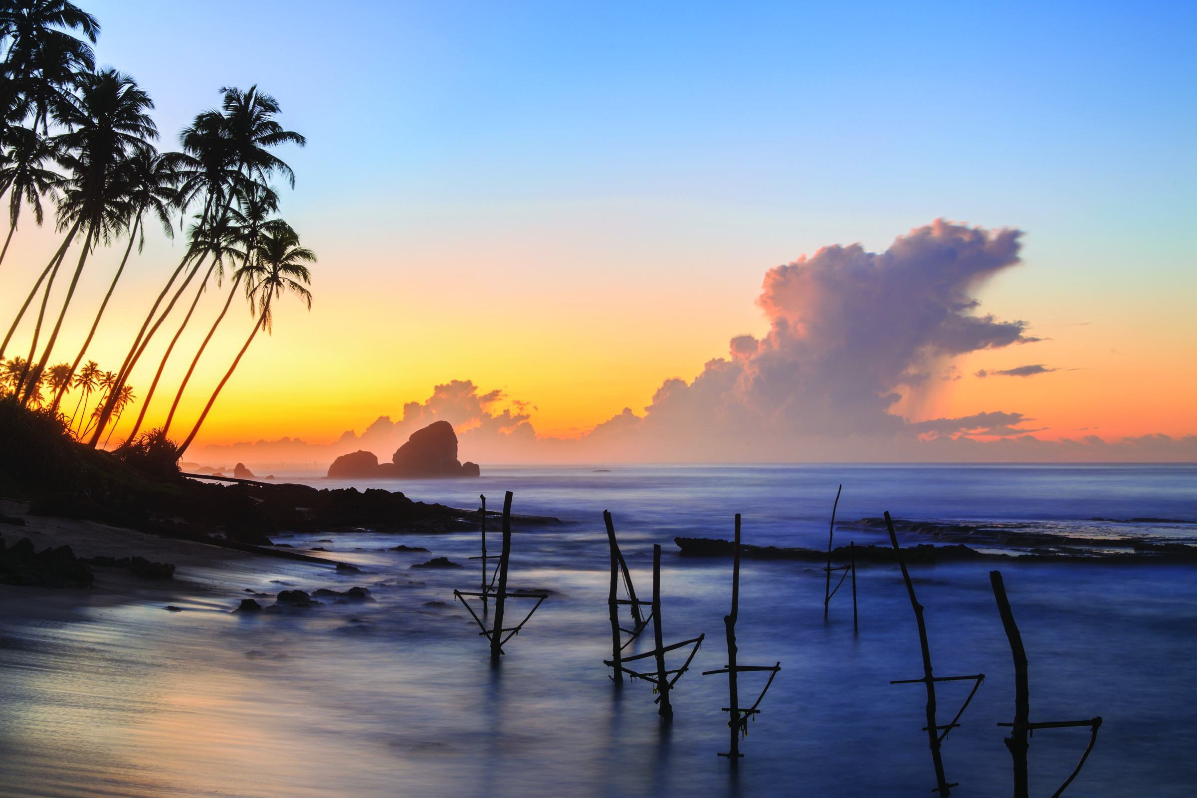 Palm trees and fishing stilts silhouetted against a colourful coastal sky